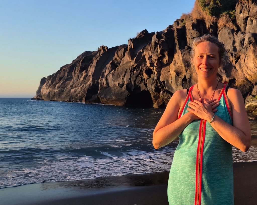 Julia Pietsch am schwarzen Sandstrand vor einer Felsküste im Abendlicht, die Hände auf dem Herzen